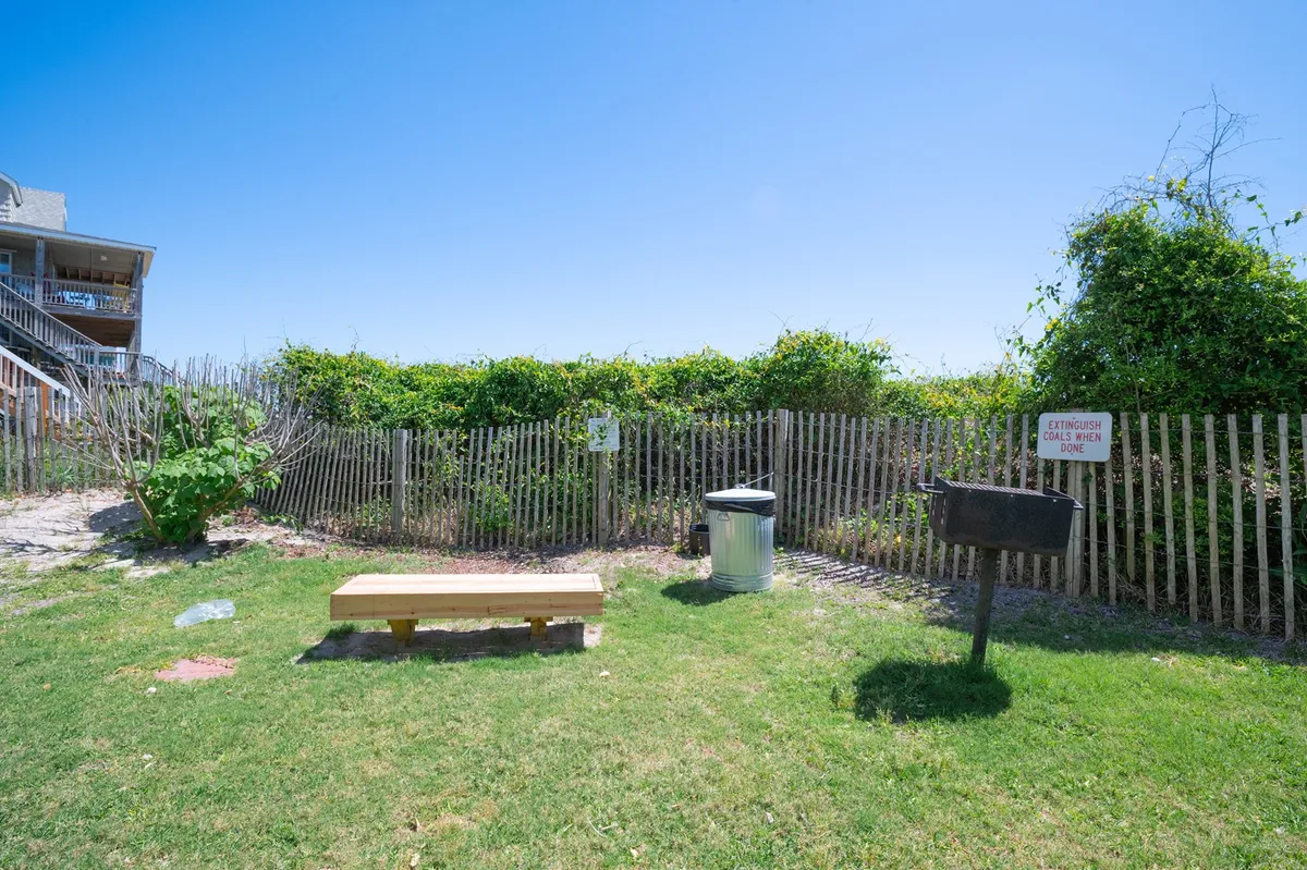 The image shows a casual outdoor area with a grill, wooden bench, green foliage, and a fence, creating a cozy spot for barbecuing, picnicking, or relaxing outdoors. The setting is inviting for community gatherings or quiet afternoons in a green space.