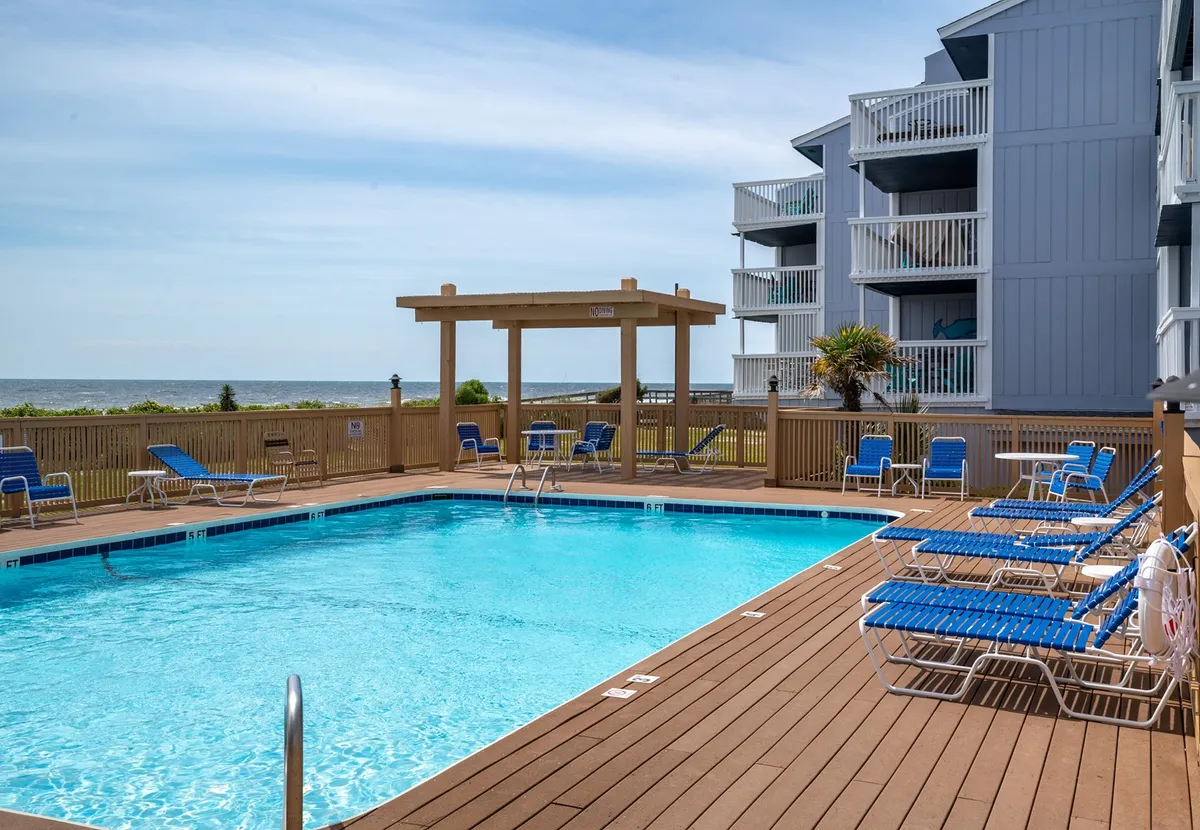 The image shows a sparkling swimming pool surrounded by inviting sun loungers and a charming pavilion, all set against the backdrop of a sweeping ocean view. The scene radiates relaxation, with guests enjoying leisurely swims, sunbathing, and socializing poolside, while the sun sets over the Atlantic. The overall mood is tranquil and welcoming, perfectly blending the comforts of the condo with the natural beauty of Carolina Beach.