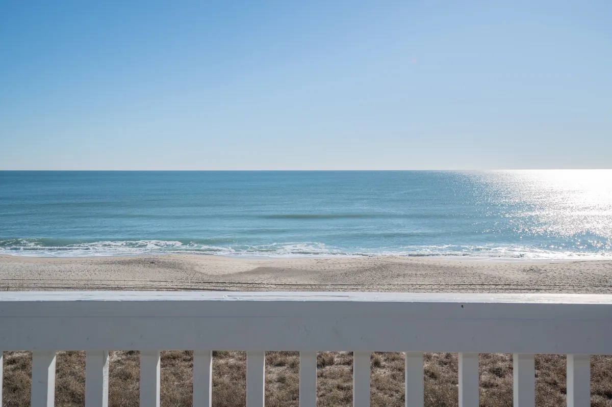 A stunning oceanfront view with the vast sand beach stretching out to the horizon, bordered by a simple railing that suggests this shot is from a spacious balcony. The scene captures gentle waves, open sky, and a laid-back coastal atmosphere, offering a glimpse of the inviting seaside just steps away.
