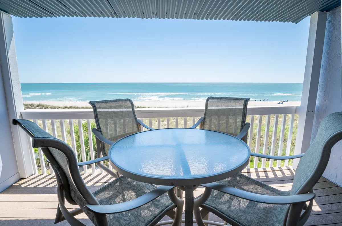 An exterior shot of the spacious balcony, featuring a round glass-top table and four comfortable chairs facing a sweeping ocean view. The scene conveys tranquil mornings, social afternoons, and leisurely sunset dinners, with the Atlantic waves as a backdrop.