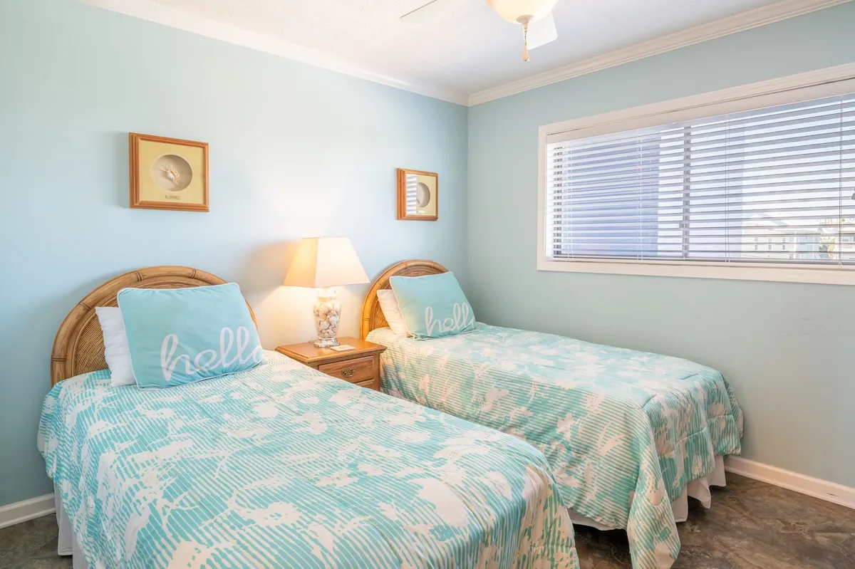 A guest bedroom with two twin beds dressed in aqua bedding, a bedside lamp, and cheerful decorative wall art. The room feels fresh, bright, and perfect for kids or guests, encouraging both comfort and togetherness.