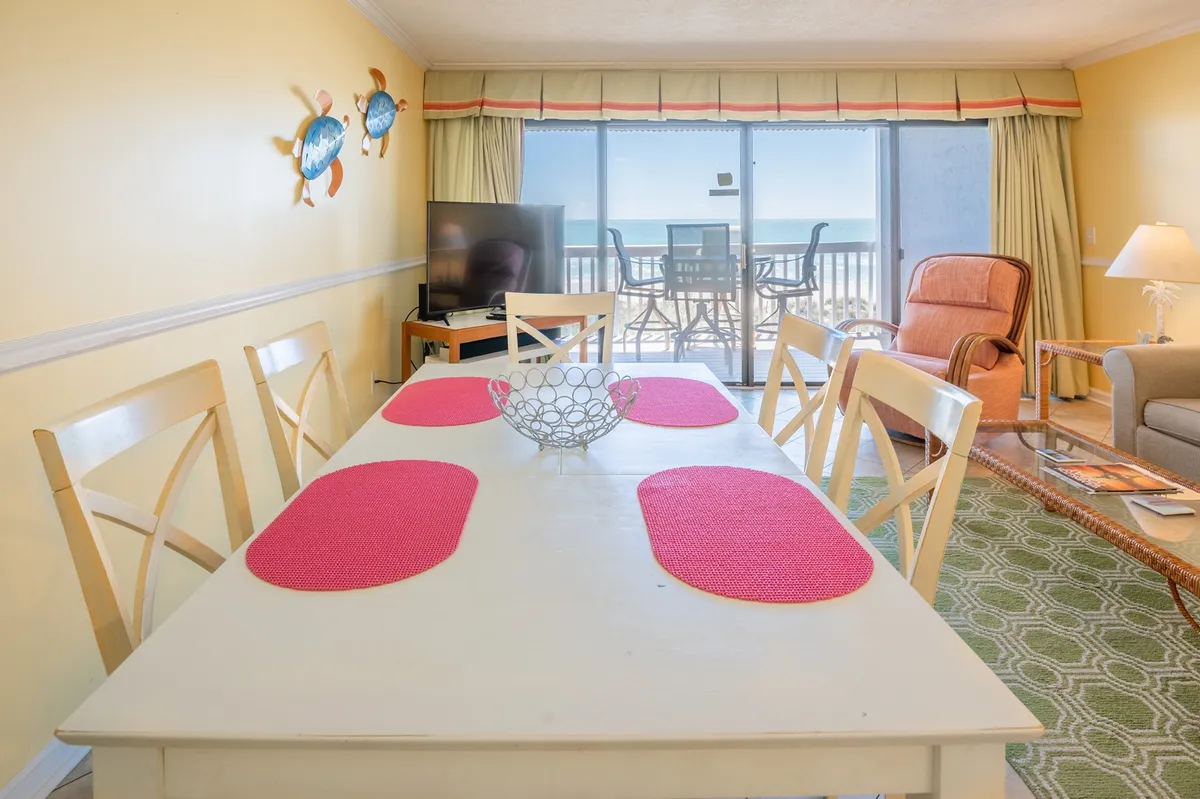 This image shows a bright and inviting dining area with a table ideally positioned to overlook the ocean through large windows. The space also includes a TV and a comfortable seating arrangement, emphasizing the unit’s open flow and suitability for family meals, game nights, or casual entertainment.