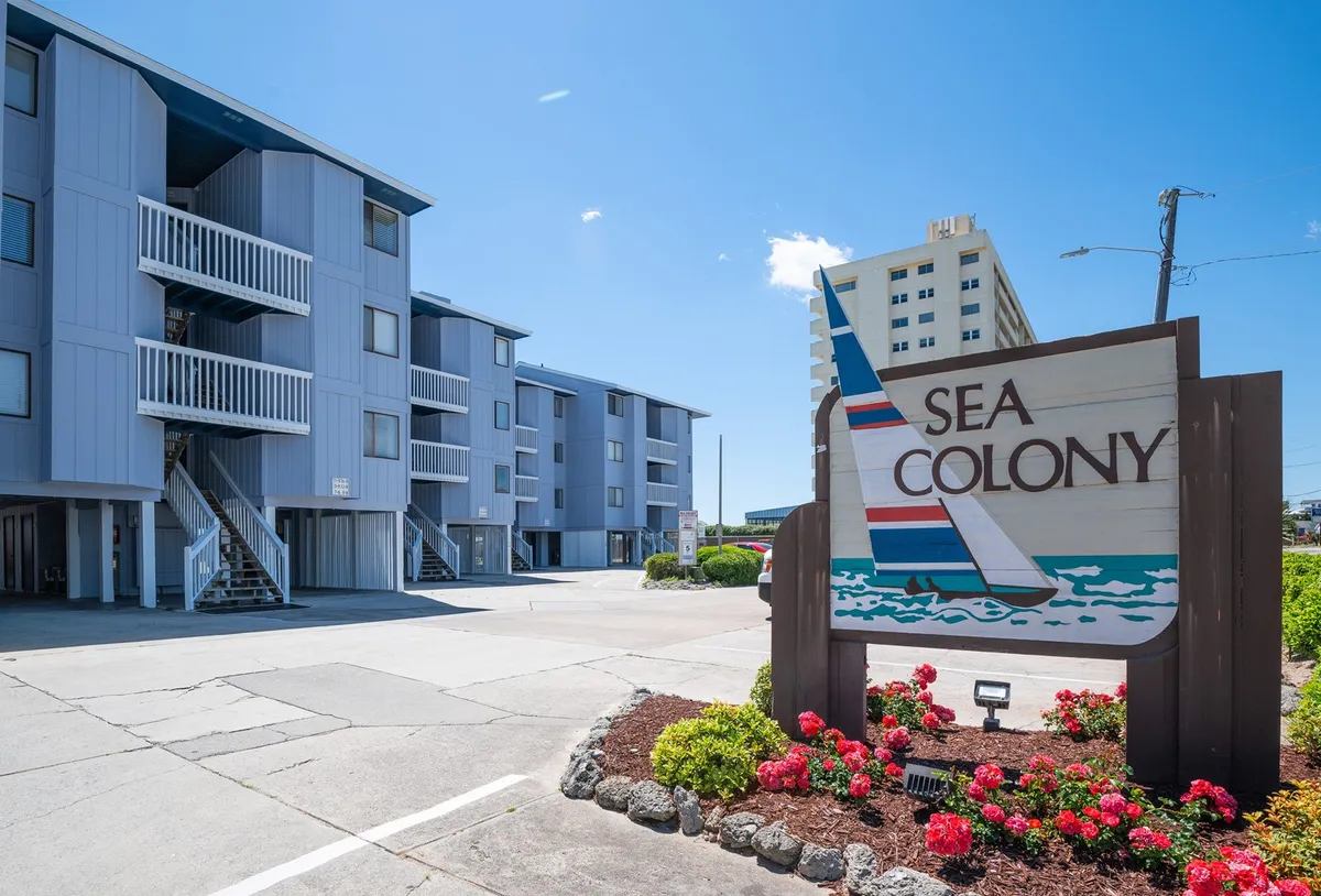 The cheerful entrance sign of Sea Colony is surrounded by colorful flowers and well-maintained landscaping, with glimpses of the condo building and sunny skies. The scene is welcoming and hints at a vibrant, community-oriented environment.