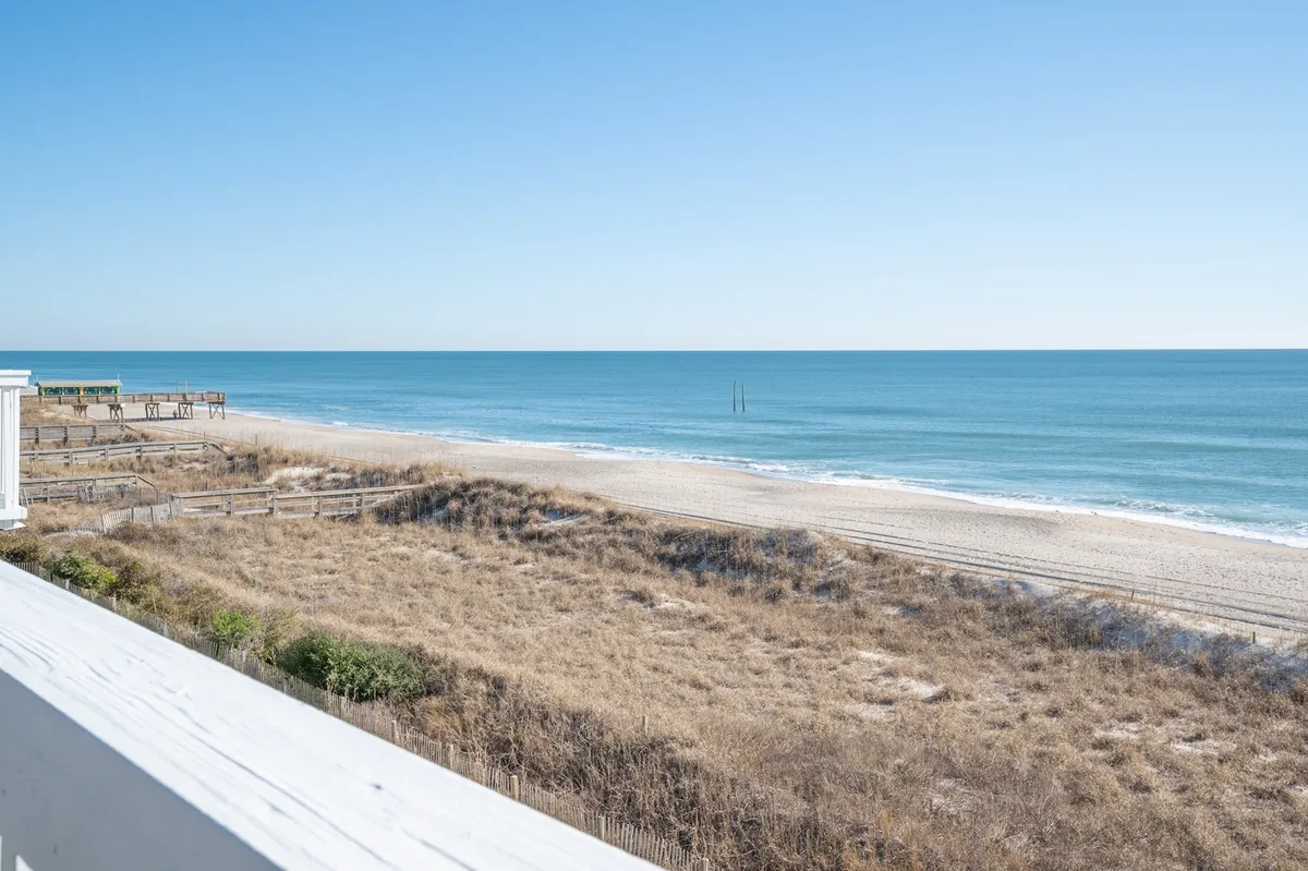A breathtaking view of the ocean, soft sandy beach, and rolling dunes stretching towards the horizon. The foreground features golden sand and gentle surf, with the vast blue sky overhead. Footprints and sandcastles might be imagined along the shore, inviting families to explore and play.